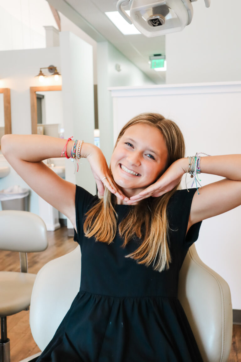 A blonde girl in a black dress showcasing her smile with her hands.
